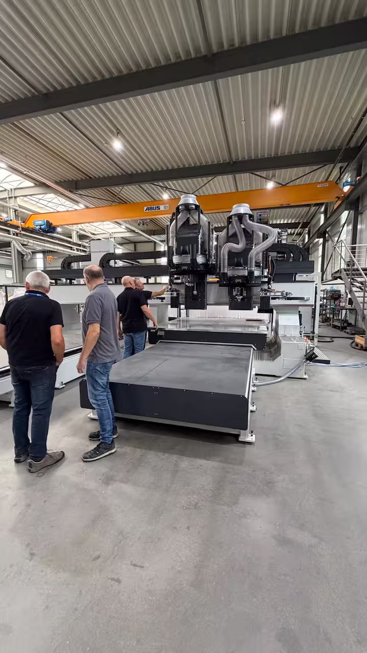 Drei Personen stehen vor einer großen CNC-Portalfräsmaschine mit zwei Fräsköpfen und Absaugschläuchen in einer Industriehalle mit ABUS-Kran