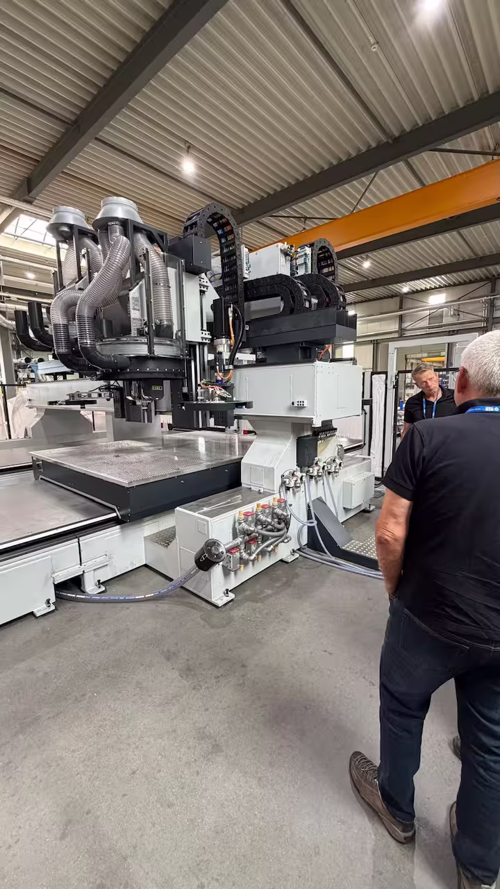 Drei Personen begutachten unsere CNC-Portalfräsmaschine mit zwei Fräsköpfen und Absaugschläuchen unter einem orangefarbenen ABUS-Hallenkran