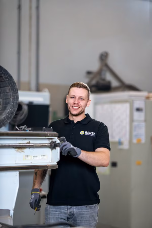 Lächelnder Mitarbeiter mit Handschuhen hält ein Bauteil an einer CNC-Maschine.