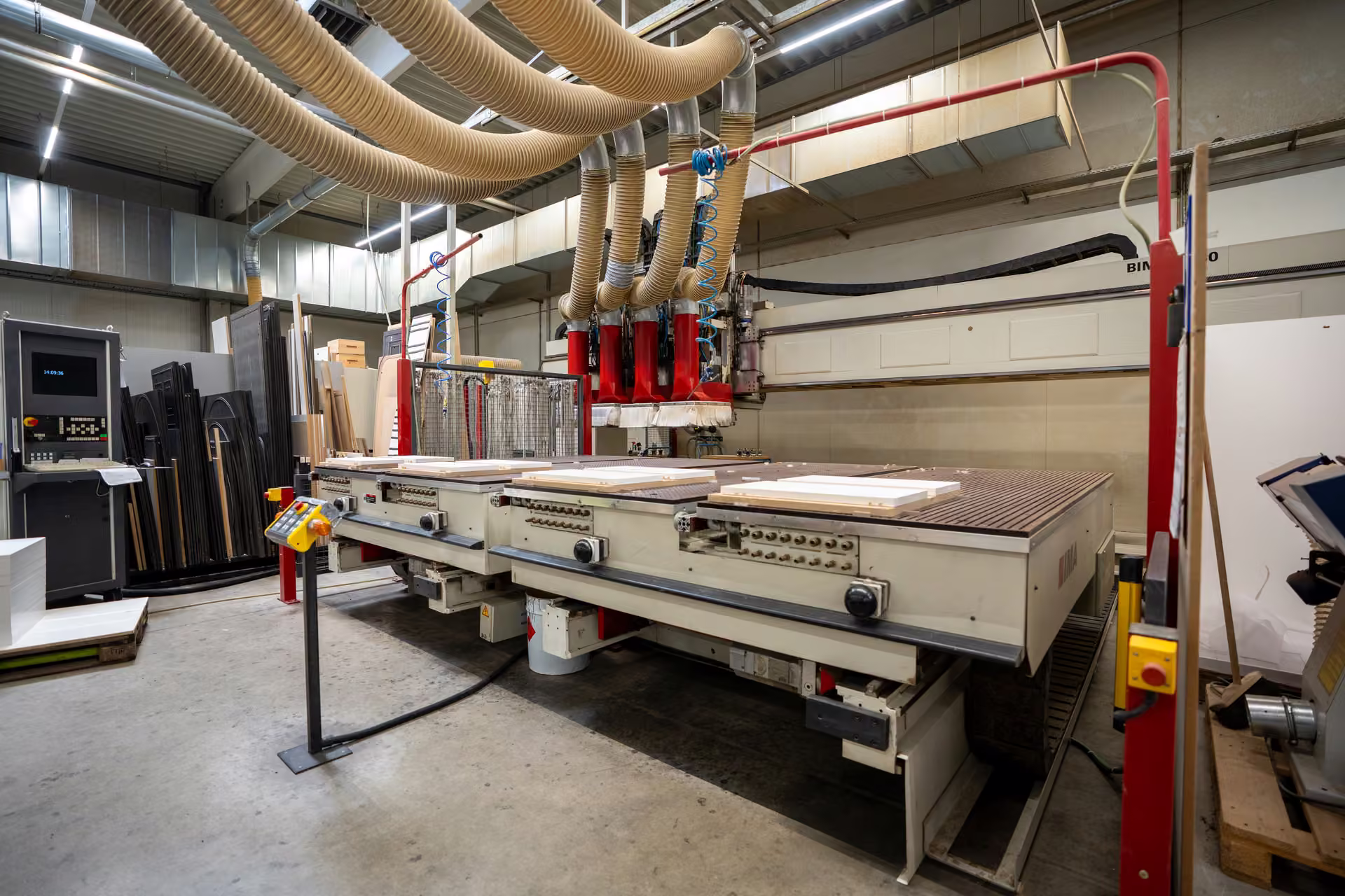 CNC-Bearbeitungszentrum mit rotem Portal und mehreren Absaugschläuchen in der Werkhalle, auf dem Maschinentisch liegen helle Werkstücke.