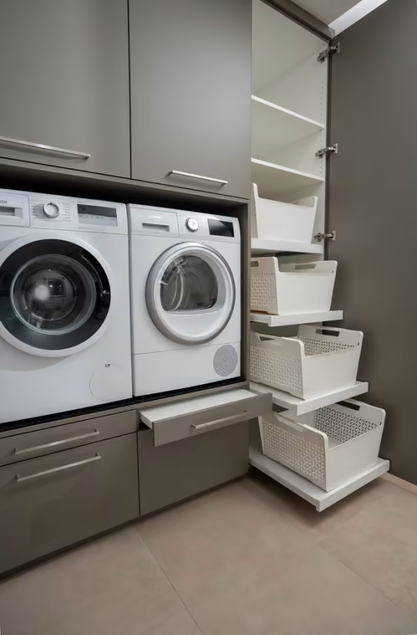 Hauswirtschaftsraum mit erhöht eingebauter Waschmaschine und Trockner, daneben offene Auszüge mit Körben.