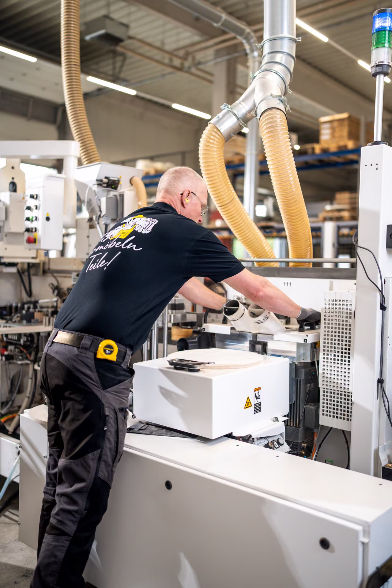 Mitarbeiter arbeitet an einem Aggregat der Kantenanleimmaschine unter gelben Absaugschläuchen, Hochformat.