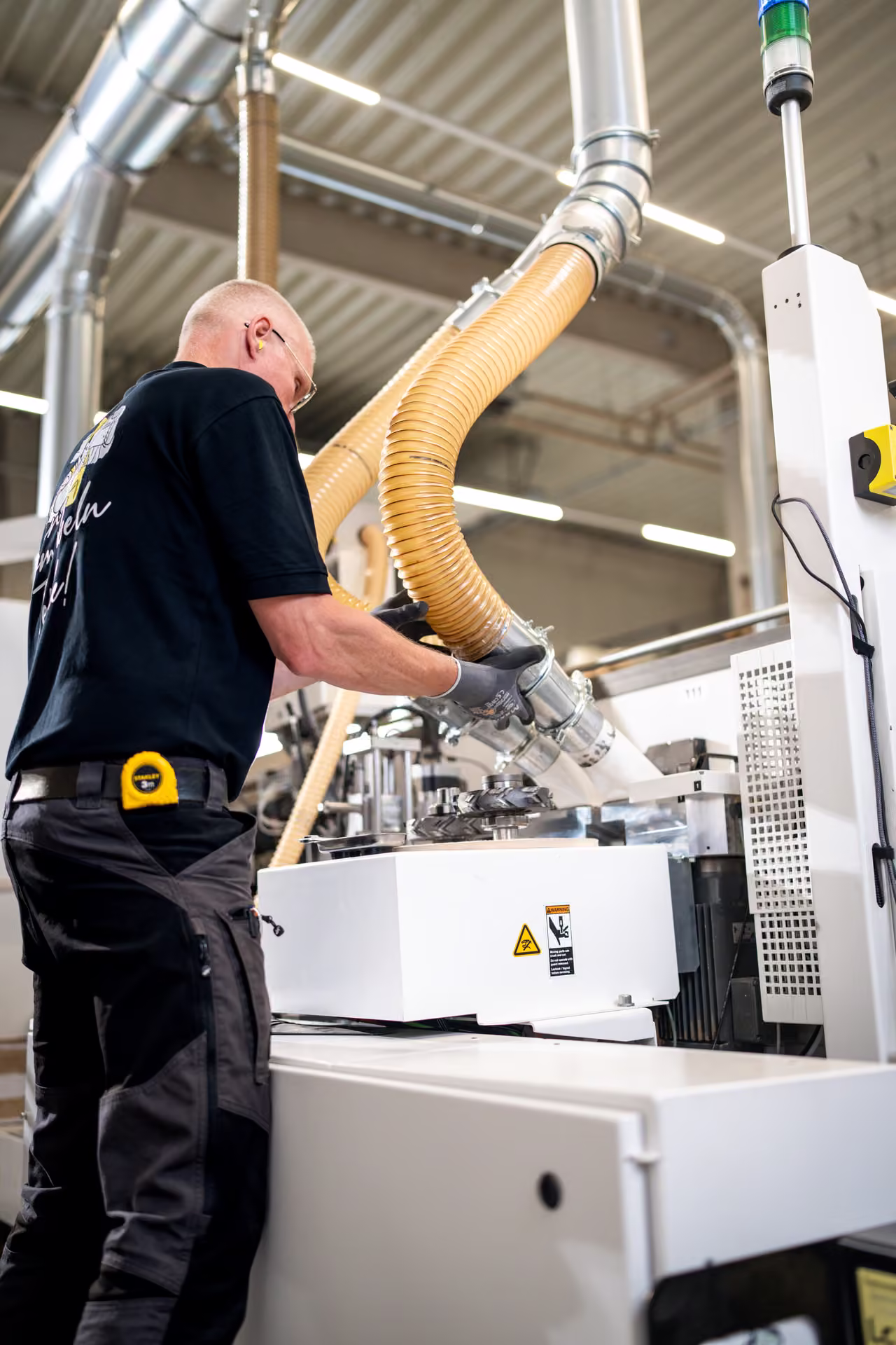 Mitarbeiter arbeitet an der Kantenanleimmaschine unter gelben Absaugschläuchen, Hochformat.