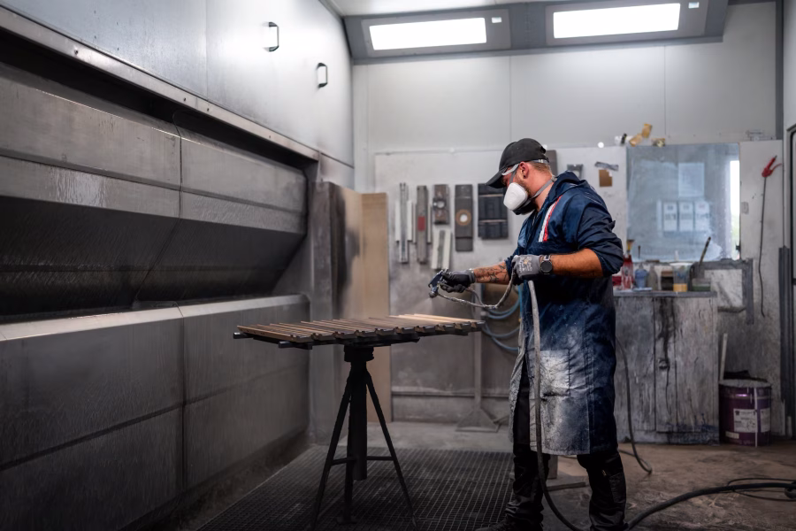 Lackierer mit Atemschutz spritzt in der Lackierkabine ein Werkstück auf einem Bock.