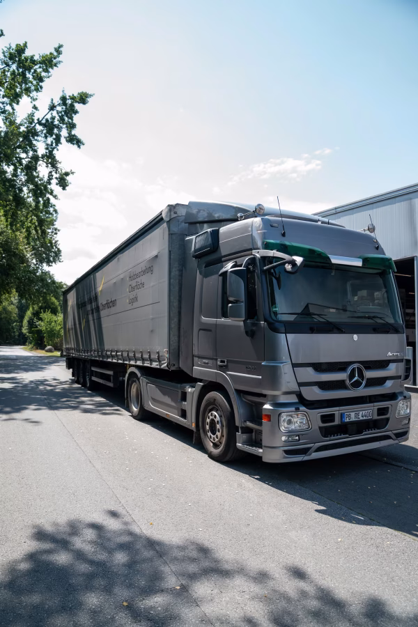 Silberner Mercedes-Benz-Sattelzug mit Planenauflieger fährt über das Werksgelände.