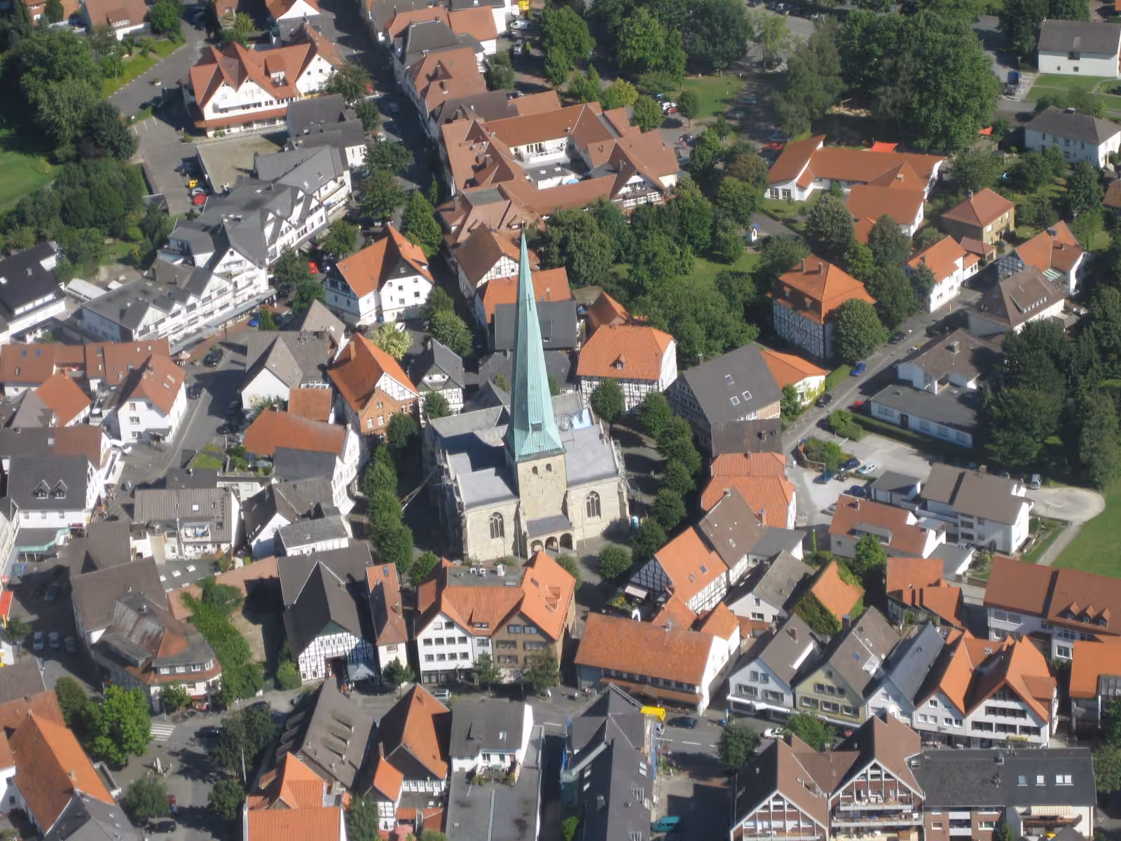 Luftaufnahme des Ortskerns von Delbrück mit Kirche und grünem Turmhelm, umgeben von Häusern mit roten Dächern und Bäumen.
