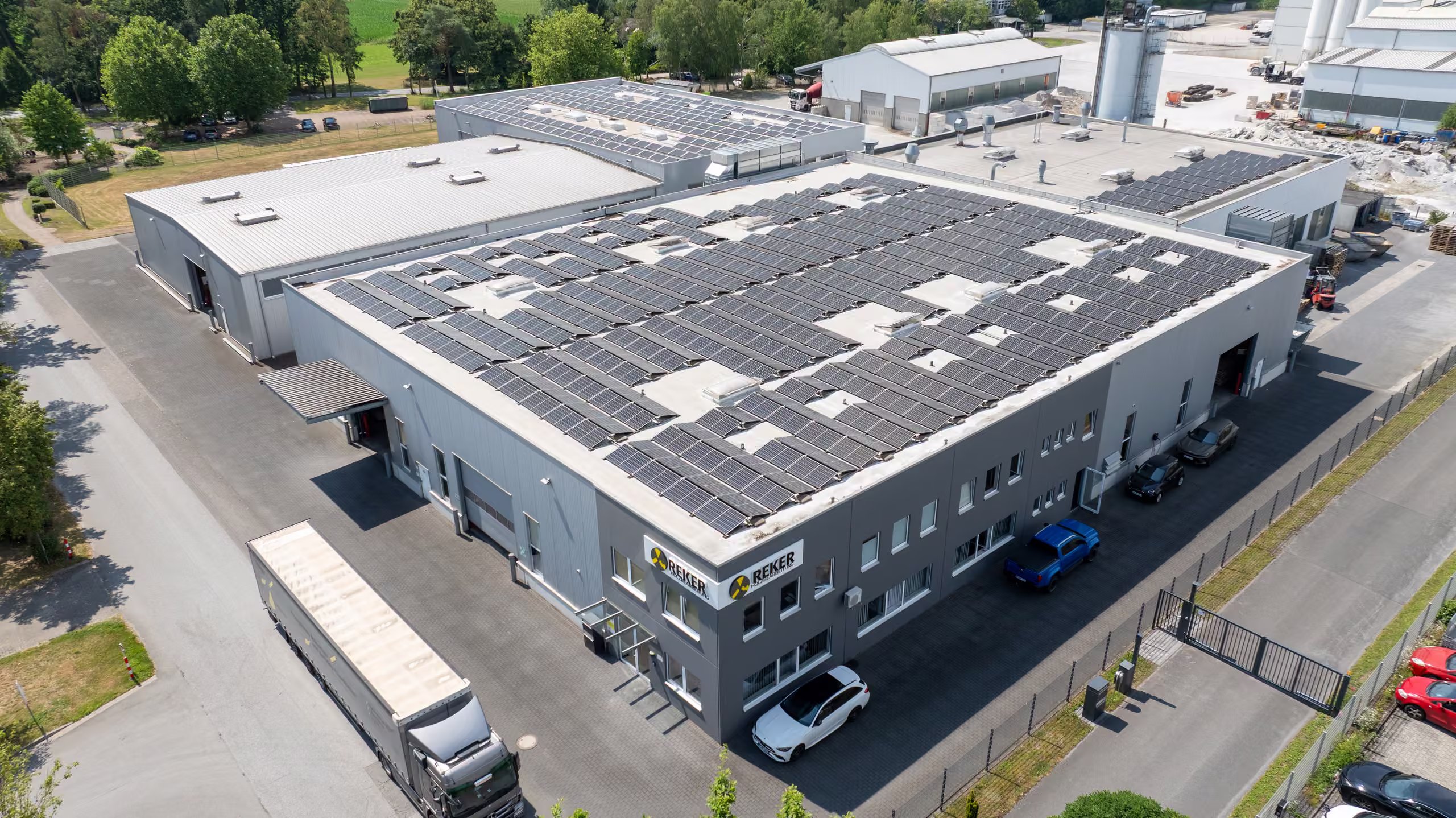 Luftaufnahme des Firmengeländes mit großen Hallen, Solaranlage auf dem Dach und Parkplätzen.