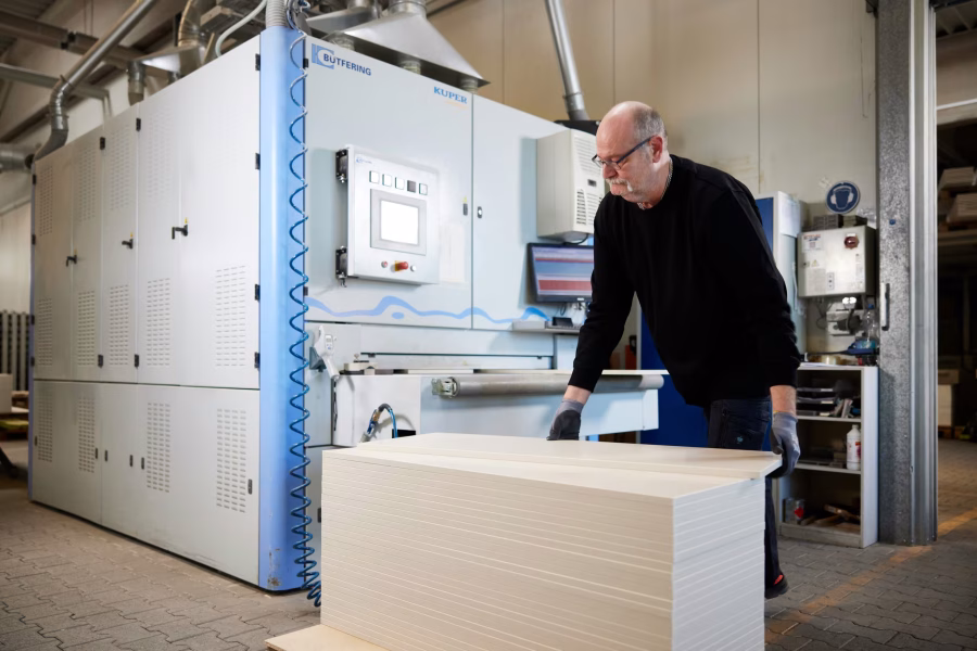 Mitarbeiter mit Brille arbeitet an einer großen Maschine, davor ein Stapel heller Platten.