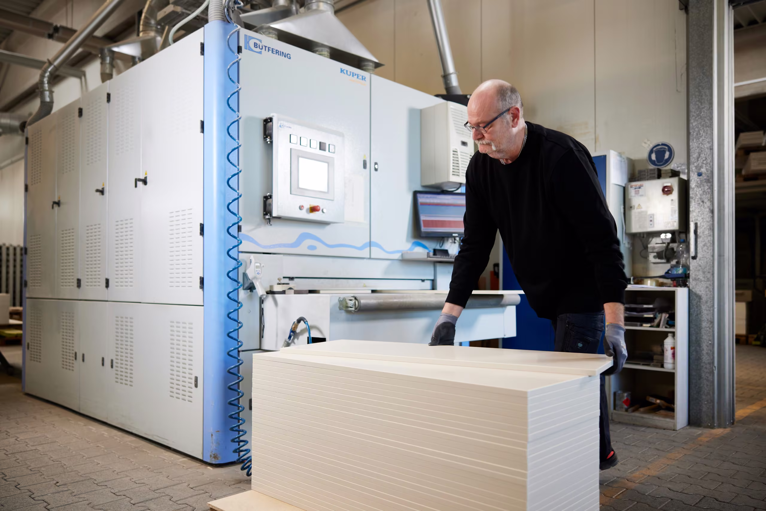 Mitarbeiter mit Brille arbeitet an einer großen Maschine, davor ein Stapel heller Platten.