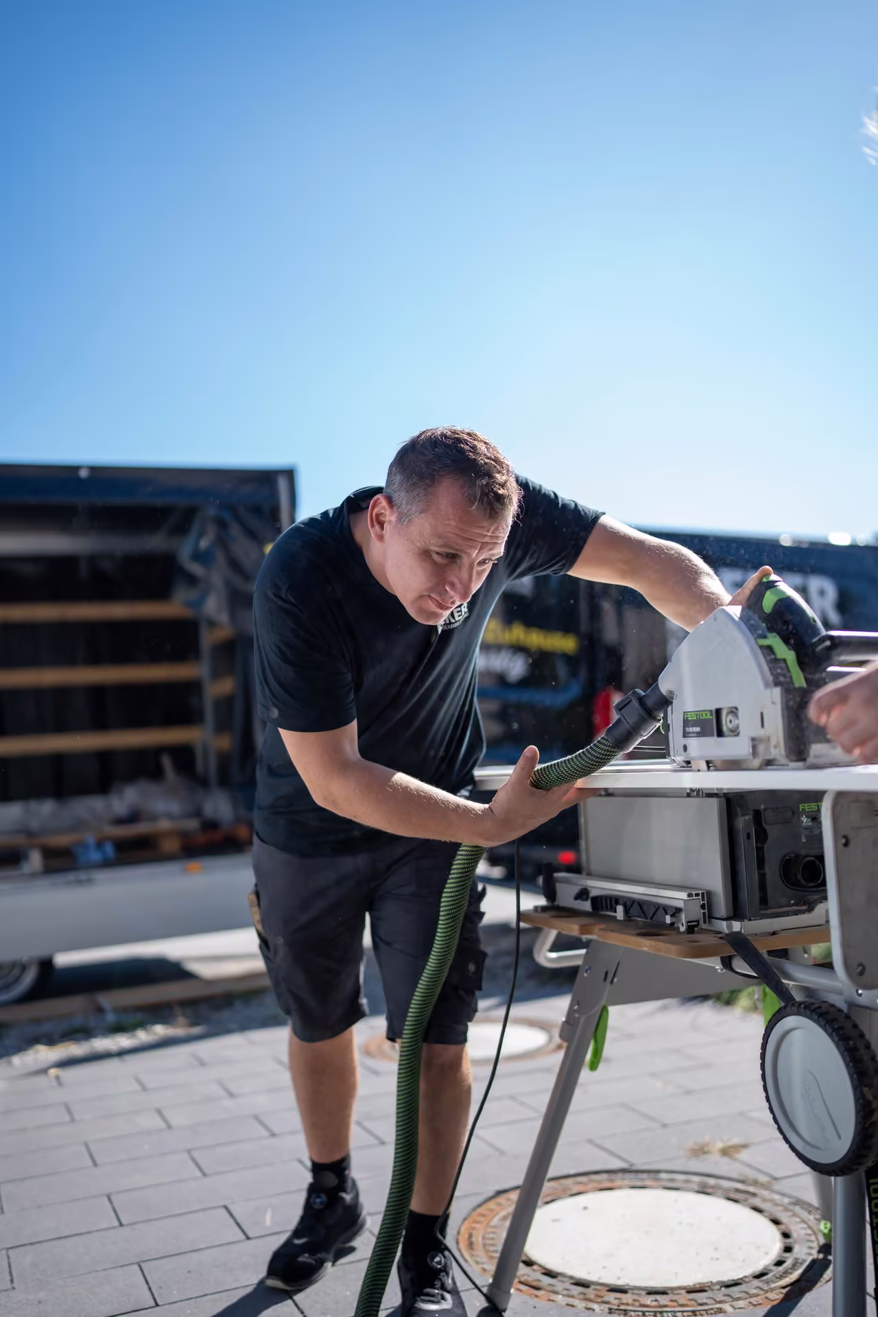 Monteur sägt vor dem offenen Transporter ein Bauteil auf einer mobilen Säge zu, Hochformat.