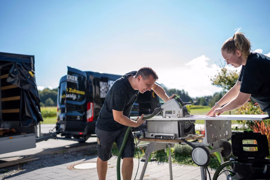 Zwei Monteure schneiden vor dem Transporter auf einer mobilen Säge ein Bauteil zu.