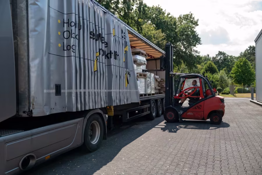 LKW-Auflieger mit geöffneter Plane auf dem Betriebshof, dahinter ein roter Gabelstapler beim Beladen.
